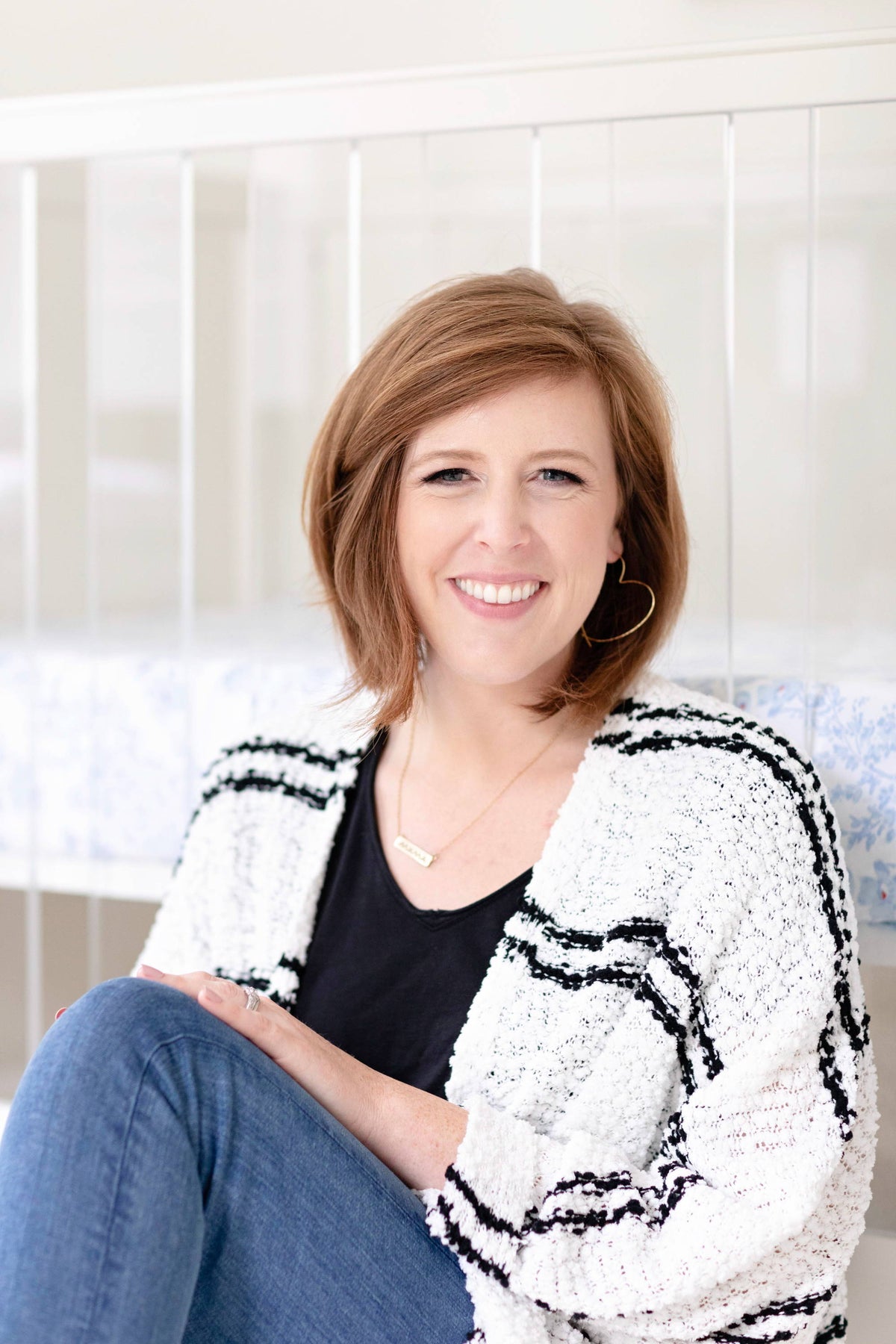 Smiling woman seated with crossed legs in a nursery, wearing a black top and textured white-and-black cardigan