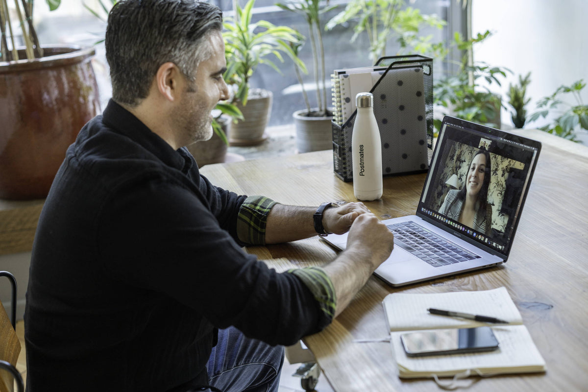 Man video calling on a laptop at a wooden desk with a notebook, phone, and white insulated bottle, plants visible through a window
