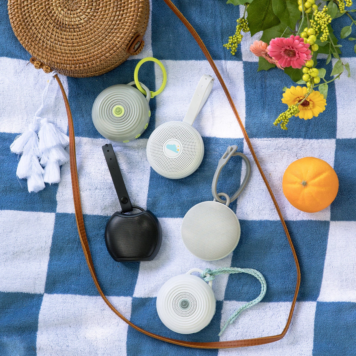 Travel sound machines arranged on a blue checkered blanket with a woven bag, flowers, and orange