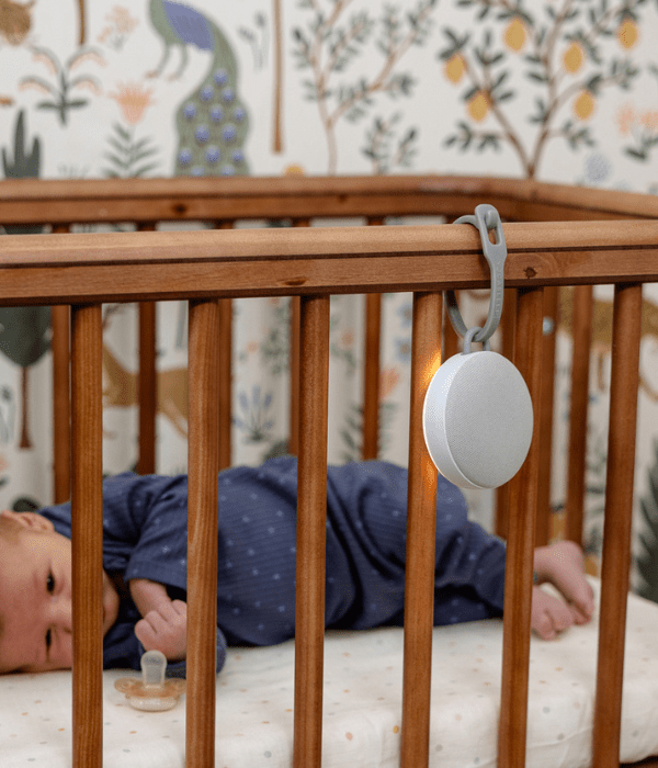 White sound machine hanging on a wooden crib beside a sleeping baby