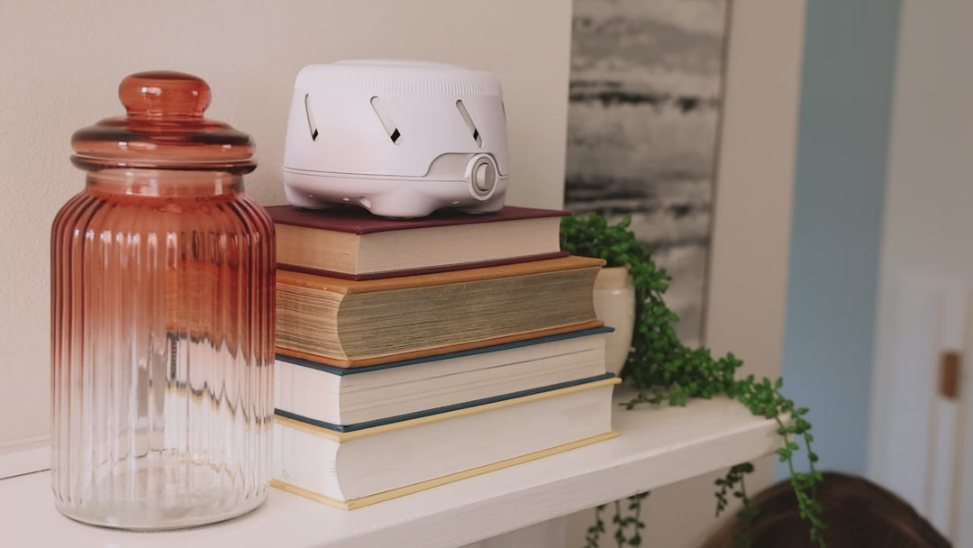 Dohm Uno white noise machine resting on a stack of books on a shelf beside a glass jar and trailing plant