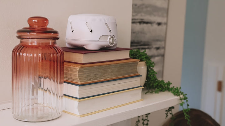 Dohm Uno white noise machine resting on a stack of books on a shelf beside a glass jar and trailing plant