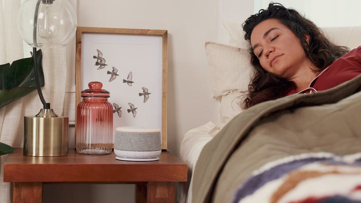 Woman sleeping as a Dohm Nova sound machine with glowing night light sits on a wooden bedside table beside a lamp and glass jar