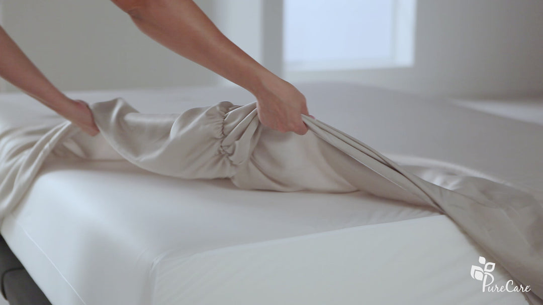 Close-up of hands fitting Premium Bamboo Sheet Set fitted sheet, showing elastic corner and smooth ivory fabric on a mattress