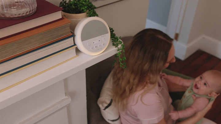 Mother holding a smiling baby beside a mantel with a Nod sound machine glowing a soft night light and visible control buttons